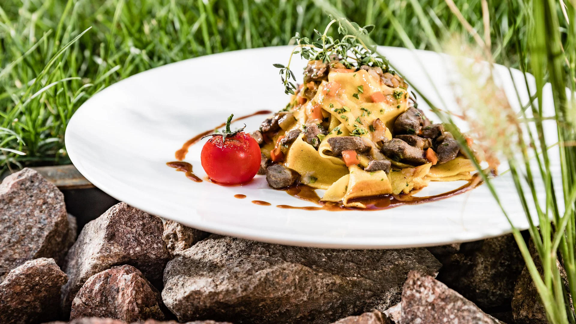 Il vostro ristorante a Termeno: cucina altoatesina e pizza Pasta con funghi e pomodoro su un piatto bianco all'aperto, appoggiato su delle pietre.